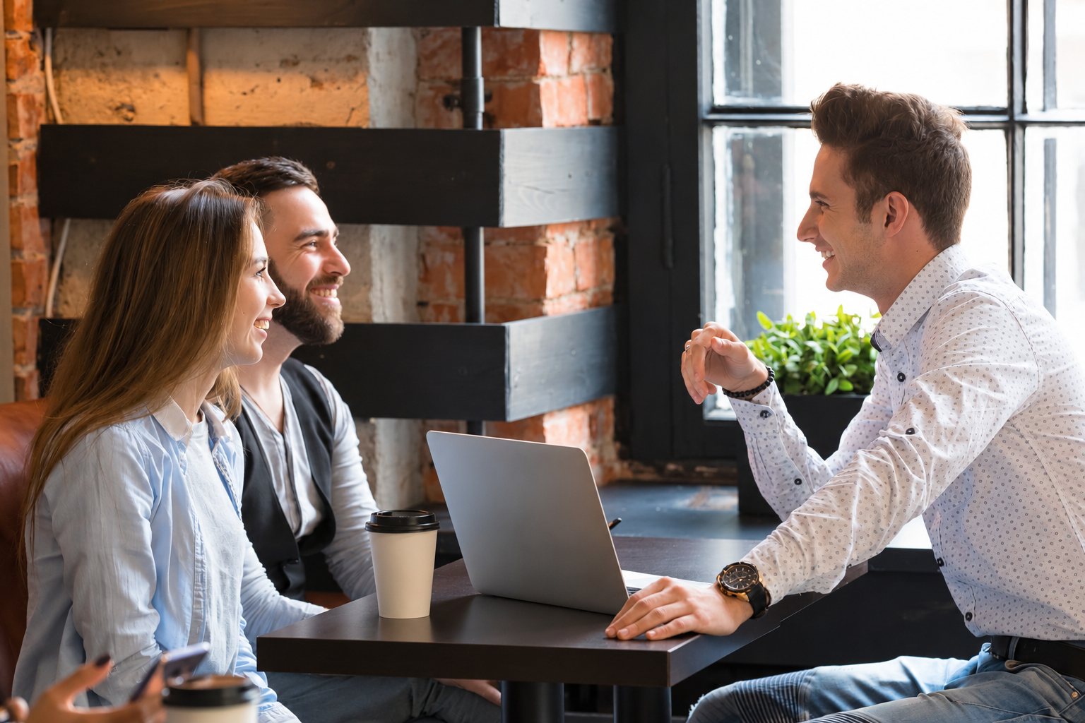 Une jeune femme, un homme barbu et un homme en chemise à motifs échangent en souriant autour d'une table avec un ordinateur portable et un gobelet à emporter, dans un café au mur de briques.