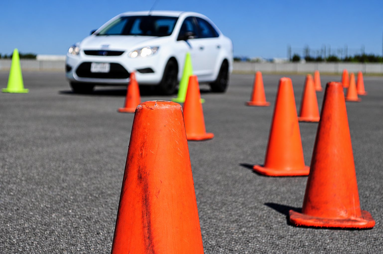 Une voiture blanche avance sur une piste en bitume entre des rangées de cônes orange et verts, avec un cône orange net au premier plan.